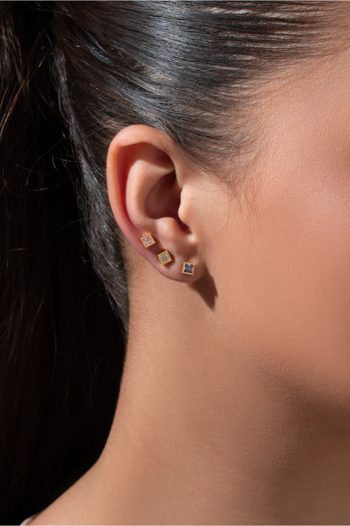 Close-up de orelha com três brincos pequenos quadrados, com pedras em tons variados de azul, dourado e rosé, usados em diferentes furos. easychic