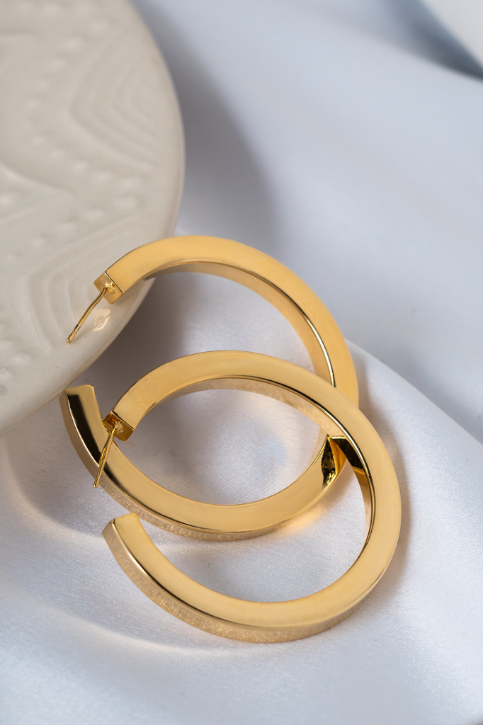 Par de brincos de argola média em acabamento dourado polido, dispostos sobre tecido acetinado branco, com detalhe de prato decorativo ao fundo. easychic
