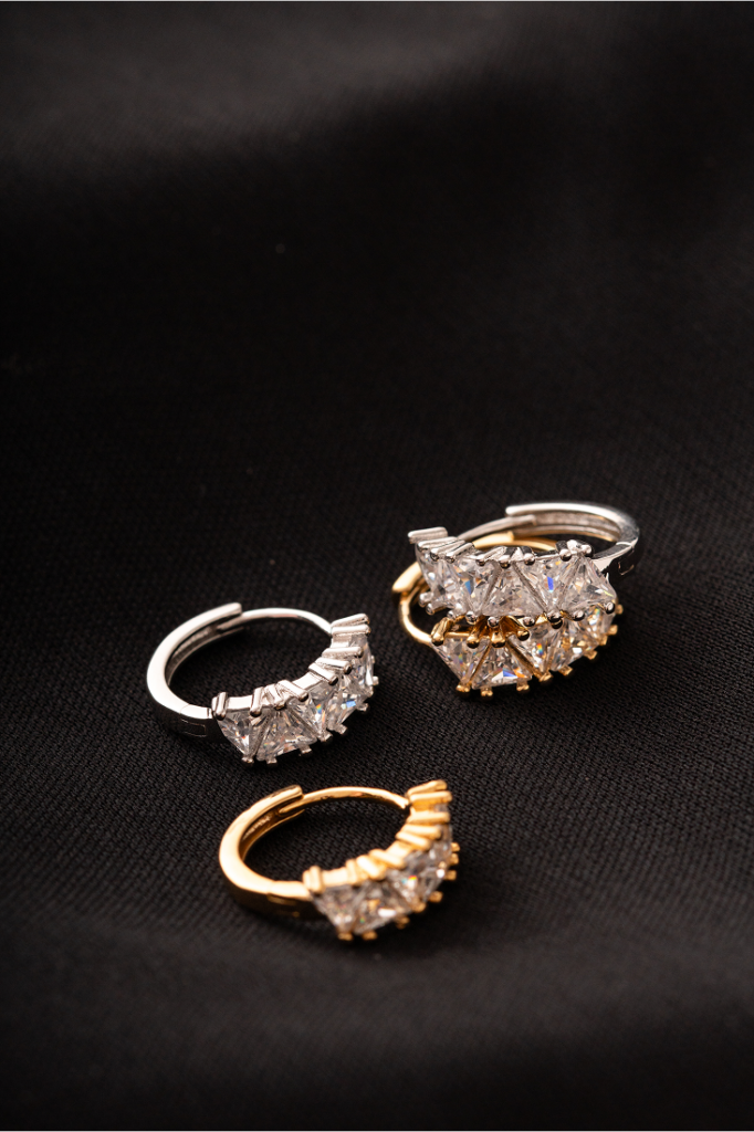Brincos pequenos de argola cravejados com zircônias em lapidação triangular, nos tons prata e dourado, dispostos sobre tecido preto. easychic