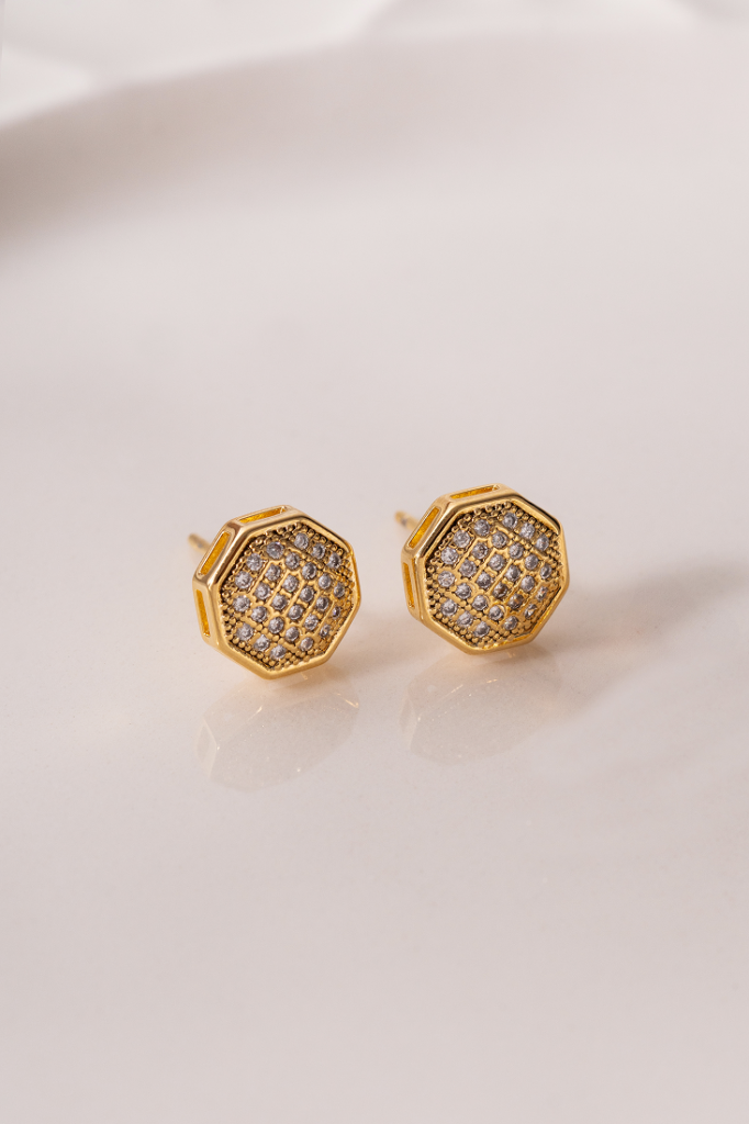 Par de brincos pequenos em formato octogonal dourado com aplicação de pedras brilhantes, apoiados sobre fundo claro. easychic