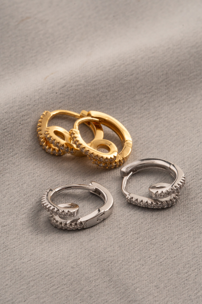 Conjunto de brincos de argola em dourado e prateado com aplicação de zircônias, dispostos sobre tecido bege claro. easychic