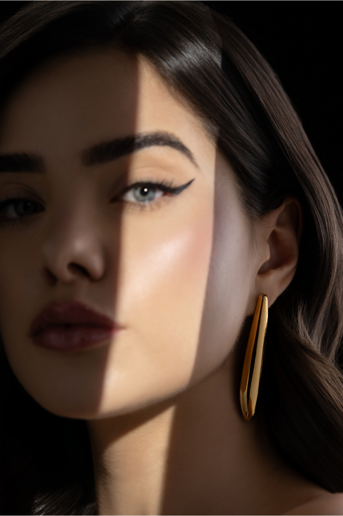Mulher com cabelo castanho e maquiagem marcada usando maxi brinco dourado com design alongado, em composição com luz e sombra no rosto. easychic